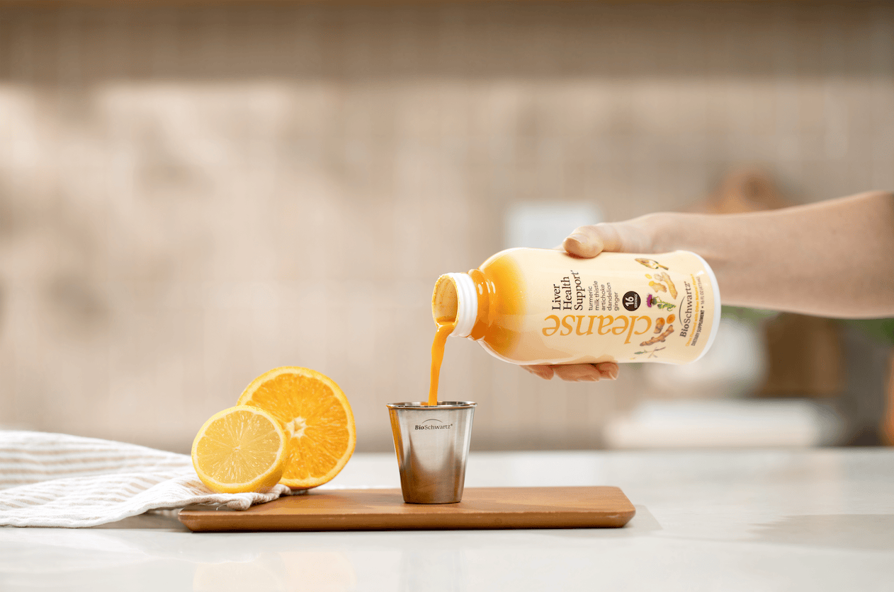 A hand pours Cleanse Liver Health Support into a metal shot glass on a wooden board, with a sliced orange and lemon beside it in a bright kitchen setting.